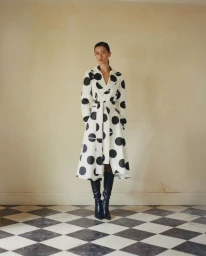 Model wearing a white polka dot trench coat and tall black boots standing on a checkered marble floor.