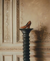 Textured brown high heel pump displayed on a black spiral wooden pedestal against an ornate classical carved wall.