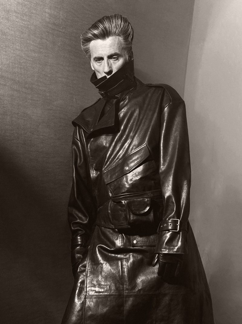 A man in a shiny, dark leather coat stands indoors against a neutral backdrop in a portrait with high contrast lighting.