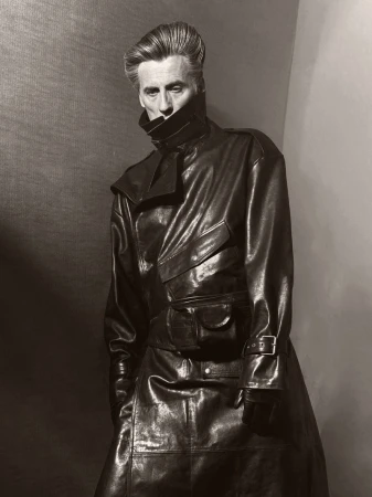 A man in a shiny, dark leather coat stands indoors against a neutral backdrop in a portrait with high contrast lighting.
