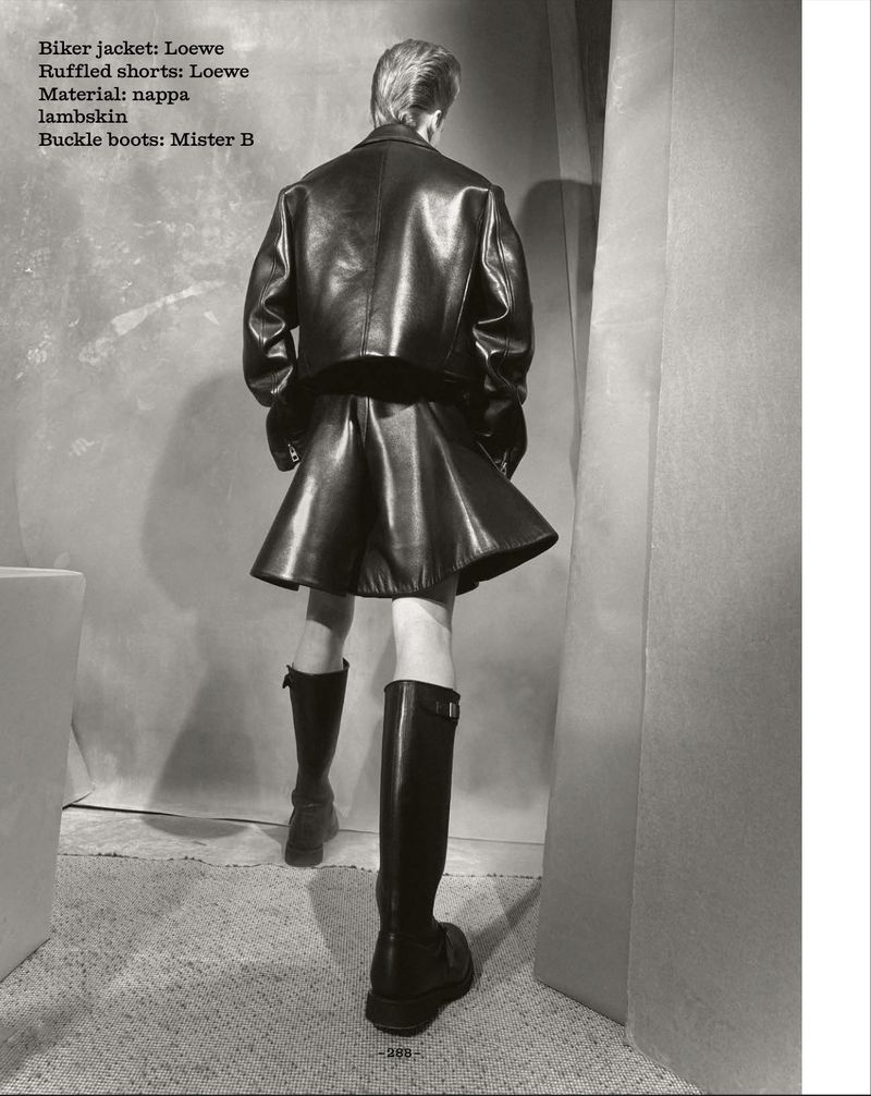 Fashion shoot of a slender person from the back wearing a black leather jacket, ruffled shorts, knee-high buckle boots, and short blond hair.
