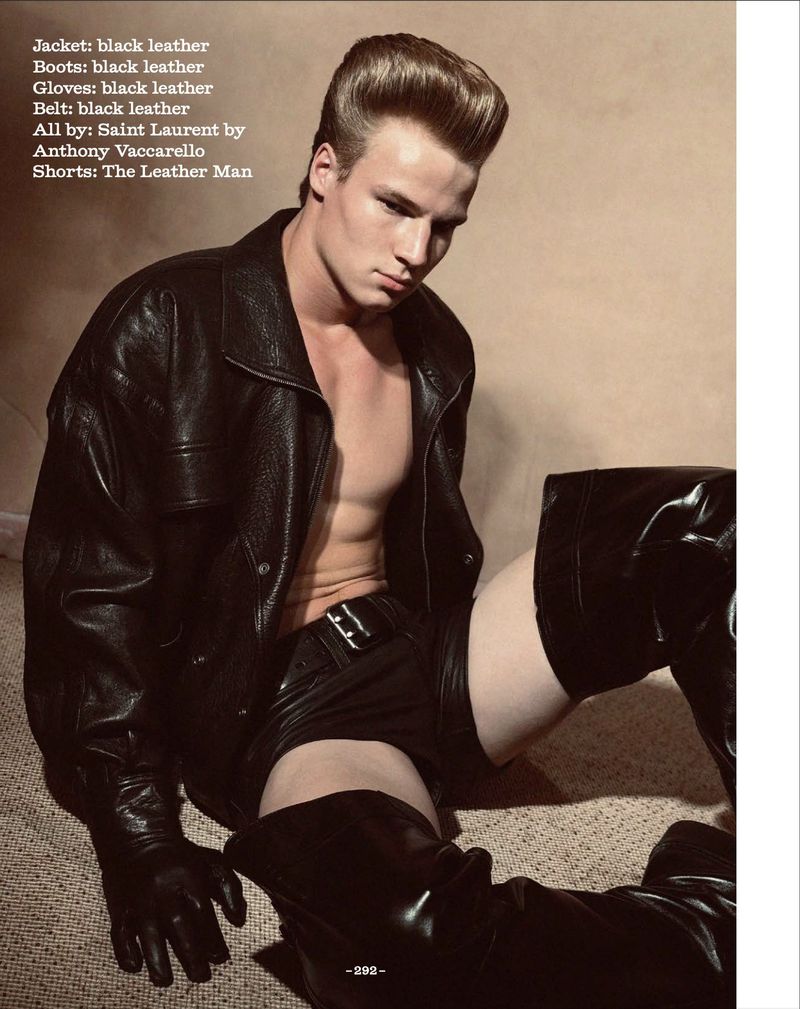 A single adult male poses in a black leather jacket, shorts, gloves, and boots against a beige background. The shot is medium.