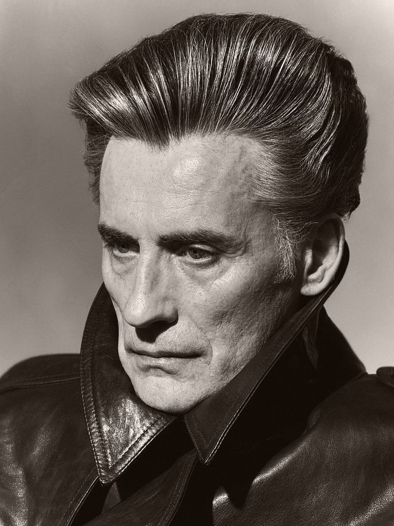 Close-up portrait of a mature man with slicked-back hair wearing a dark leather coat, set against a neutral background in a studio.
