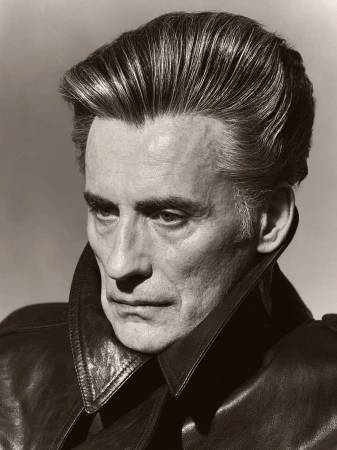 Close-up portrait of a mature man with slicked-back hair wearing a dark leather coat, set against a neutral background in a studio.