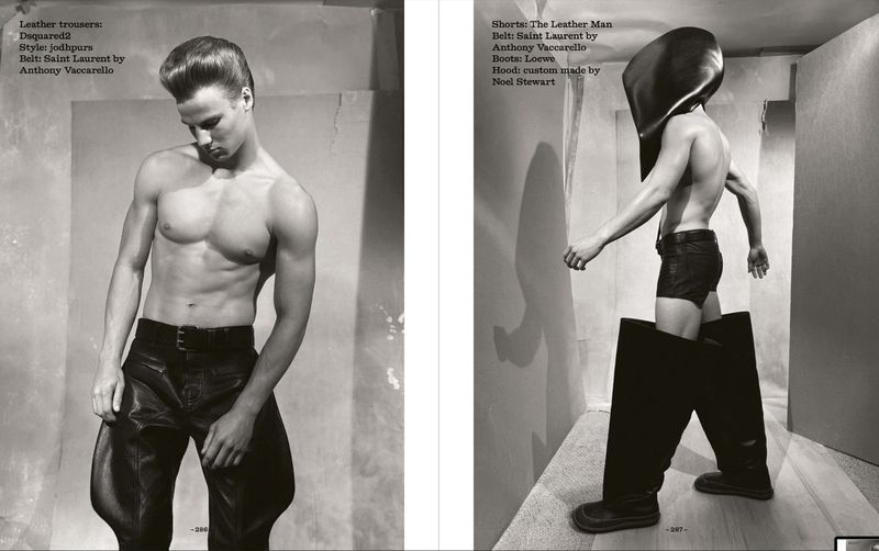 Black and white fashion shot of two shirtless male models wearing leather trousers, shorts, belts, boots, and headgear in a studio.