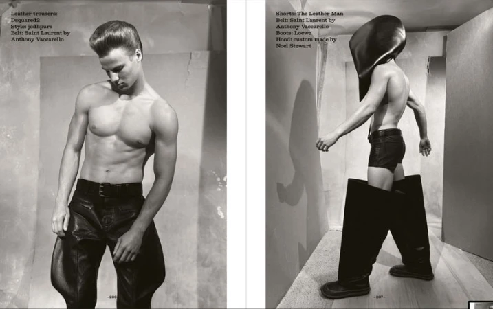 Black and white fashion shot of two shirtless male models wearing leather trousers, shorts, belts, boots, and headgear in a studio.
