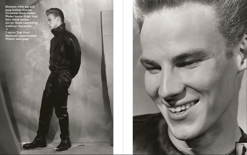 Editorial fashion shot of a blonde male model in full black leather outfit, including jacket, trousers, and boots, with a gray backdrop.