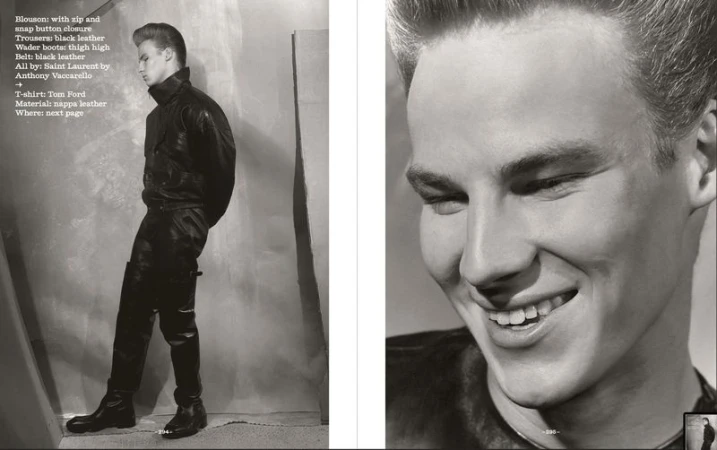 Editorial fashion shot of a blonde male model in full black leather outfit, including jacket, trousers, and boots, with a gray backdrop.