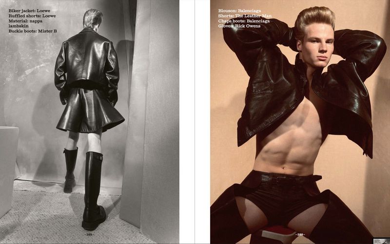 Editorial fashion shot: two shirtless male models pose indoors wearing black leather jackets, ruffled shorts, chaps boots, and gloves.