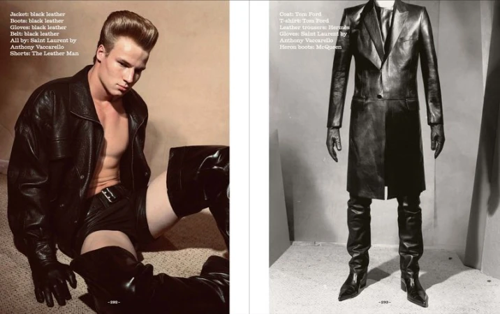 Editorial fashion shot of a male model in a black leather jacket, shorts, gloves and high boots posing indoors on a carpet with neutral wall.