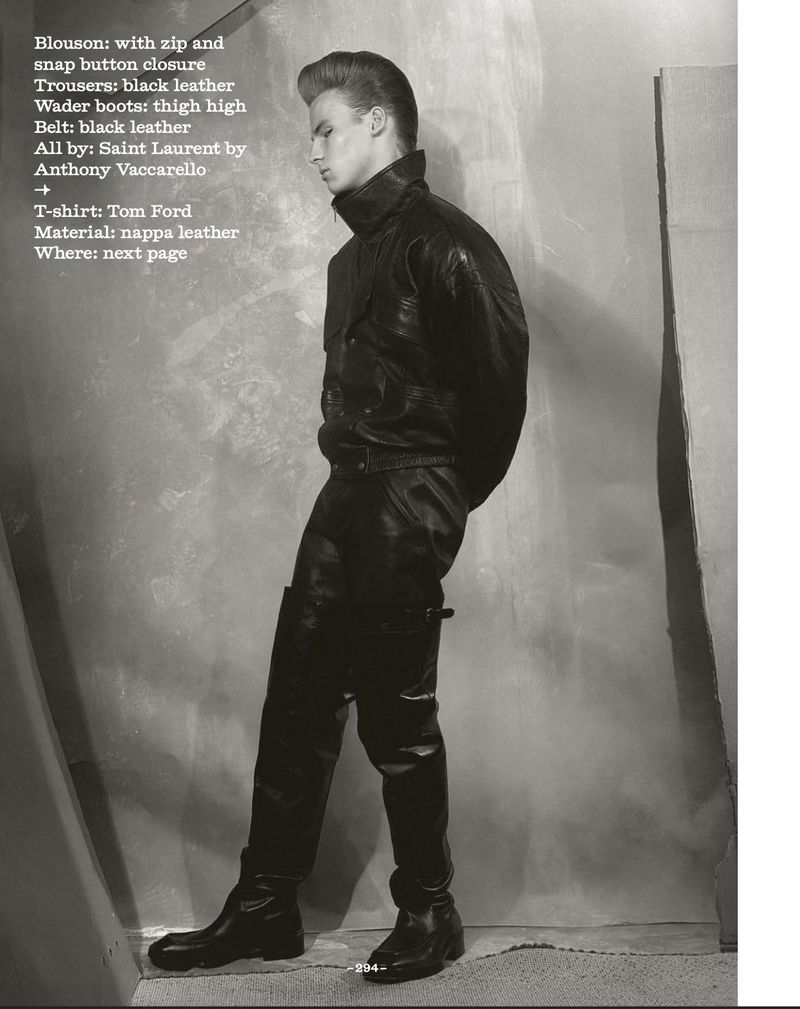 Fashion shoot features a blonde adult male model in a black leather blouson, trousers, and boots, posed in a gray studio setting.