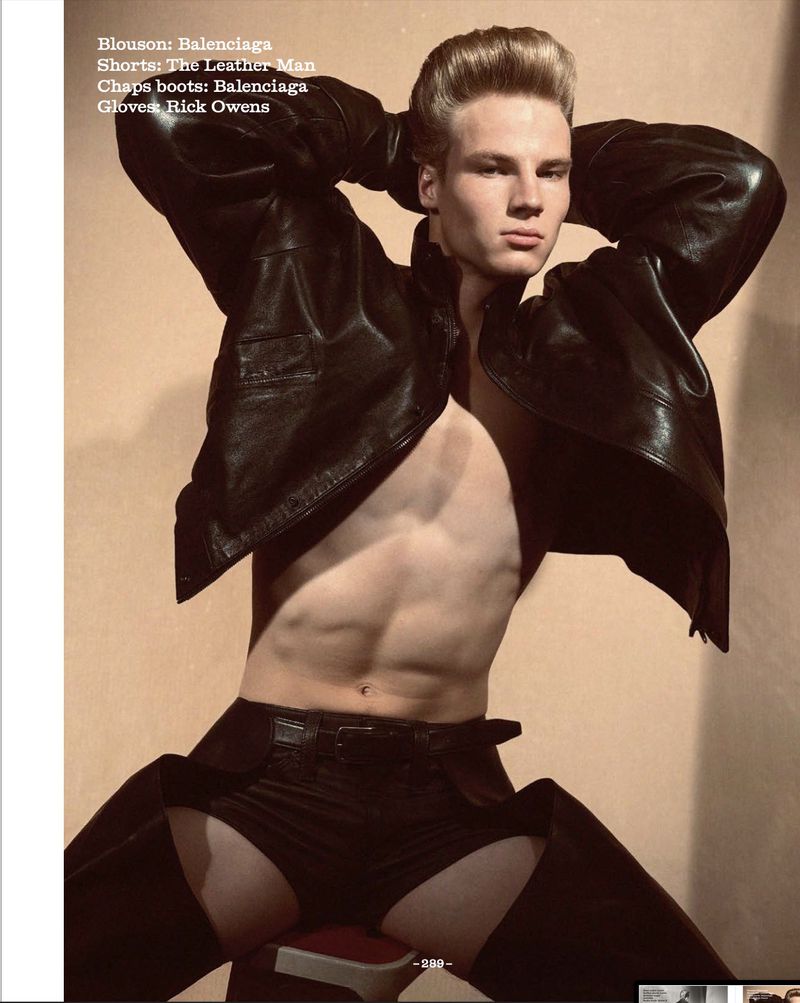 Muscular male model with pompadour hairstyle poses topless in a leather jacket, chaps, and gloves in a neutral-toned photo studio.