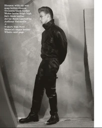 Monochrome fashion editorial of a man in a black leather blouson, trousers, and boots standing in profile against a textured studio wall.