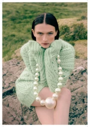Model Lara Mullen seated outdoors in an oversized mint green knit sweater wearing an exaggerated giant pearl necklace.
