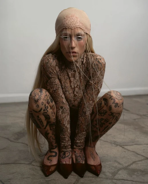 Person in crouching pose wearing lace body suit with tattoos and blonde hair. Person has light skin and is wearing red pointed shoes.