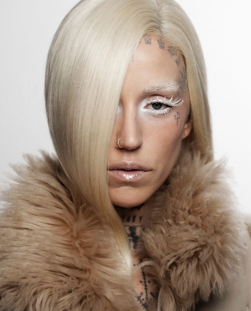 Close-up portrait of a woman with striking makeup, platinum blonde hair, and multiple face and body tattoos in a studio setting.