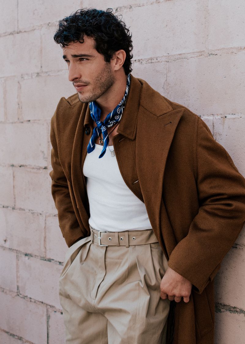 Portrait of a young adult man wearing a brown overcoat, white undershirt, paisley bandana, and tan pants, standing against a wall.