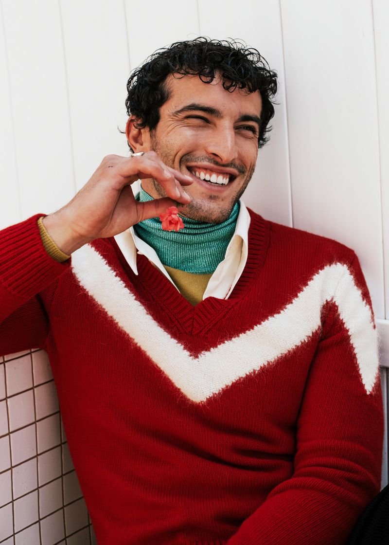 A happy man with curly dark hair smiles while holding a pink flower. He wears a V-neck sweater with a white chevron design.