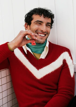 A happy man with curly dark hair smiles while holding a pink flower. He wears a V-neck sweater with a white chevron design.