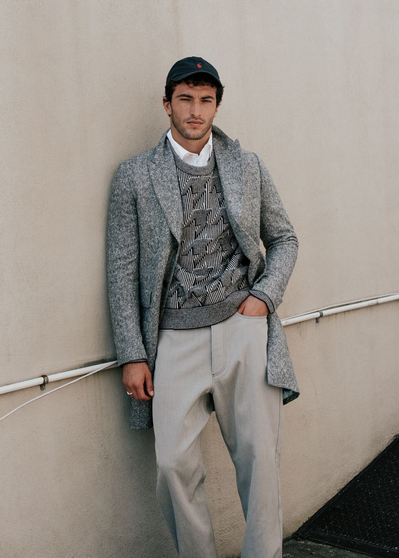 Portrait of a young adult male wearing a baseball cap, sweater, coat and slacks leaning against a wall looking at the camera.