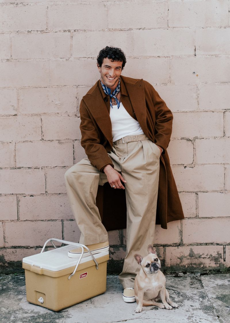 Smiling man in a brown coat poses with a French bulldog, leaning against a brick wall, with a tan cooler at his feet.