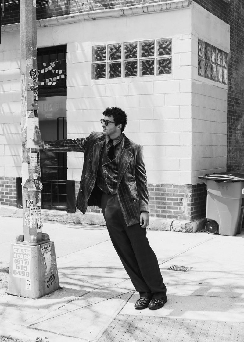 Monochrome portrait of a man in a stylish suit posing outdoors, leaning against a pole, with an urban building in the background.
