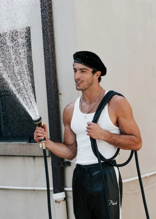 A man waters with a garden hose, smiling and wearing a black beret, white tank top, and athletic pants in the afternoon sun.