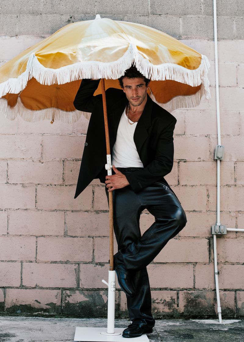 Stylish man poses outdoors under yellow umbrella. He wears a black jacket, white shirt, shiny pants, and black shoes against a brick wall.