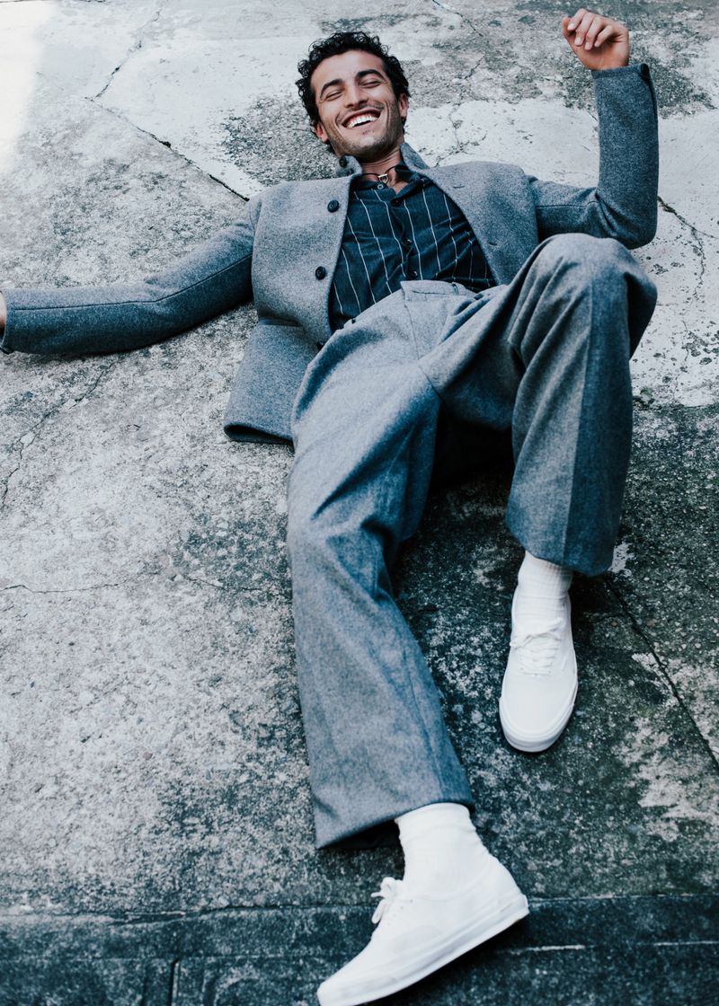 Stylish adult male in a grey suit and white sneakers reclines on concrete in an urban setting with a happy expression.