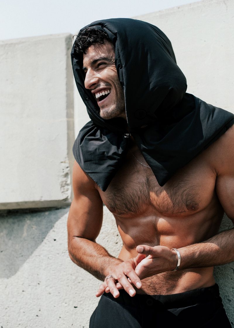 Athletic man smiles leaning against a concrete wall wearing a black hooded scarf. The subject is outdoors in bright light.