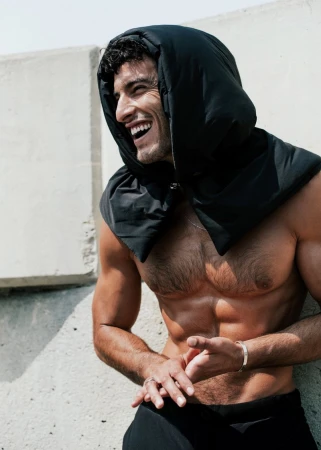 Athletic man smiles leaning against a concrete wall wearing a black hooded scarf. The subject is outdoors in bright light.