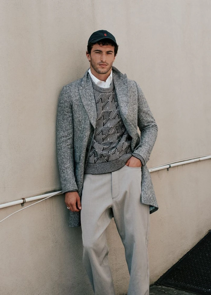 Model Raphael Diogo wearing a grey overcoat, patterned sweater, and cap leaning against a neutral wall.