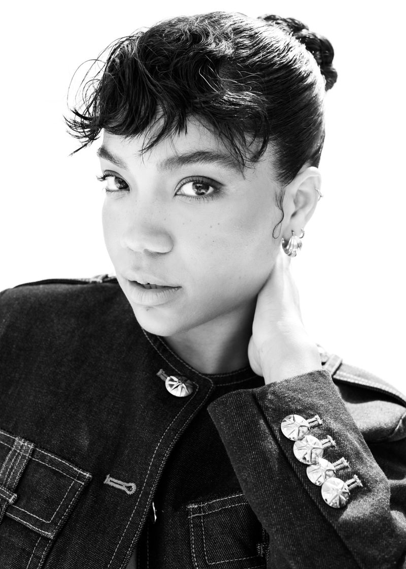 Black and white portrait of a woman in a denim jacket with silver buttons, looking at the camera against a bright white backdrop.