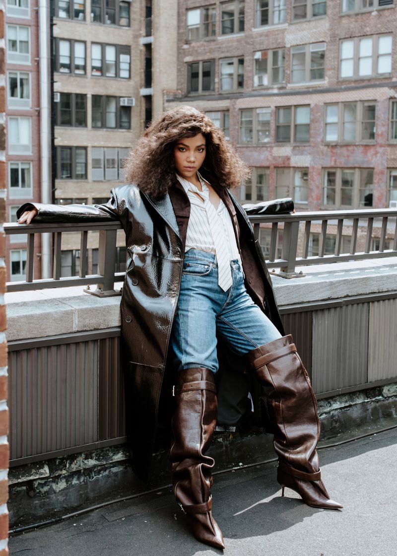 A fashionable adult woman with curly hair poses on a rooftop wearing a shiny brown coat, blue jeans, and tall brown boots in an urban setting.