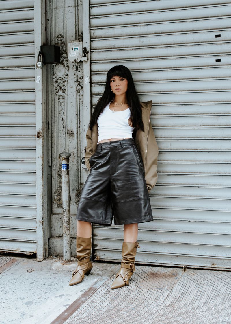 A woman in leather culottes, boots, and a jacket poses against a corrugated metal wall in urban setting, showcasing a modern fashion style.