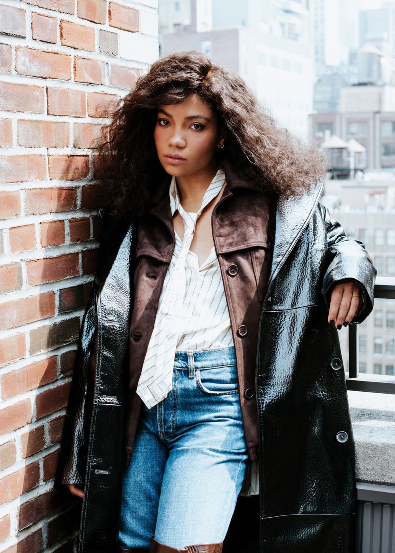 Young woman with curly hair poses outdoors in a long black coat, blue jean shorts, striped shirt, and brown jacket in an urban setting.