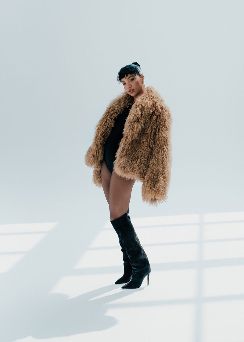 Woman in a furry coat and knee-high boots poses in front of a white background with shadow patterns. Fashion photography.