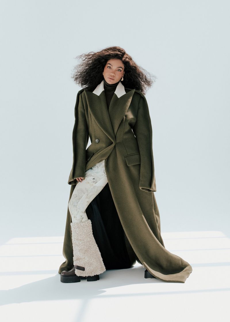 Elegant woman with curly dark hair poses in an olive green overcoat against a pale blue background in a fashion photography shot.