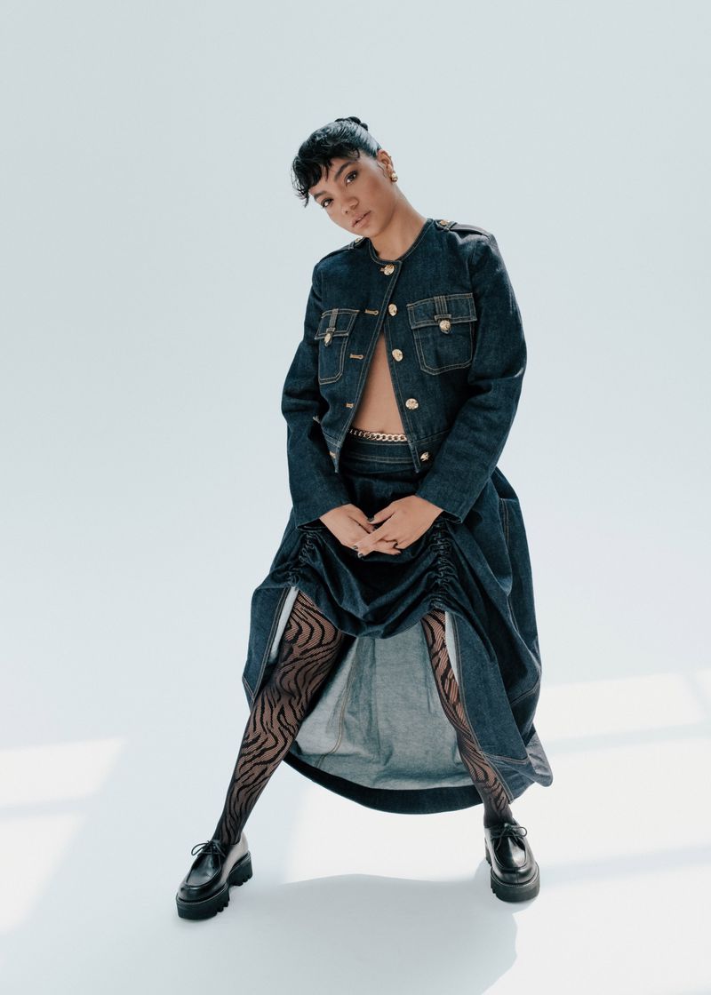 Fashionable young adult woman posing in a denim jacket, gathered midi skirt, patterned tights, and platform loafers against a white backdrop.