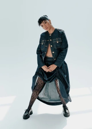 Fashionable young adult woman posing in a denim jacket, gathered midi skirt, patterned tights, and platform loafers against a white backdrop.