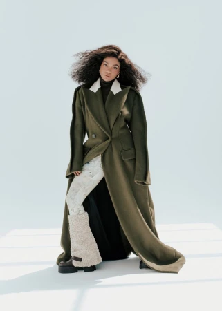Silvia Dionicio wearing a long olive green coat and shearling boots against a seamless white studio backdrop.