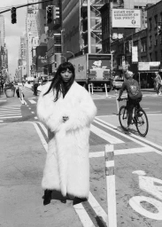 Woman in a long white fur coat standing on a busy city street intersection in a monochrome fashion portrait.