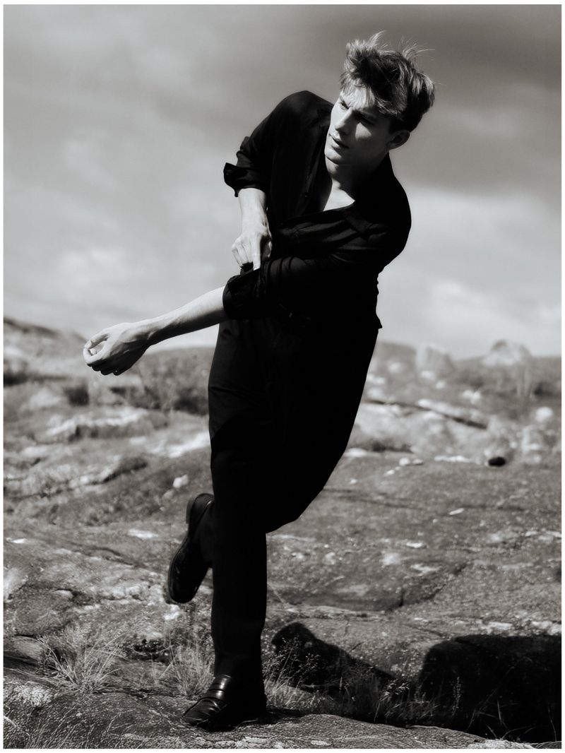 Monochrome outdoor shot of a young adult man in a dark shirt and trousers rolling up his sleeve against a cloudy sky and rocky terrain.