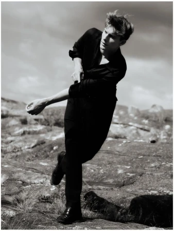 Monochrome outdoor shot of a young adult man in a dark shirt and trousers rolling up his sleeve against a cloudy sky and rocky terrain.