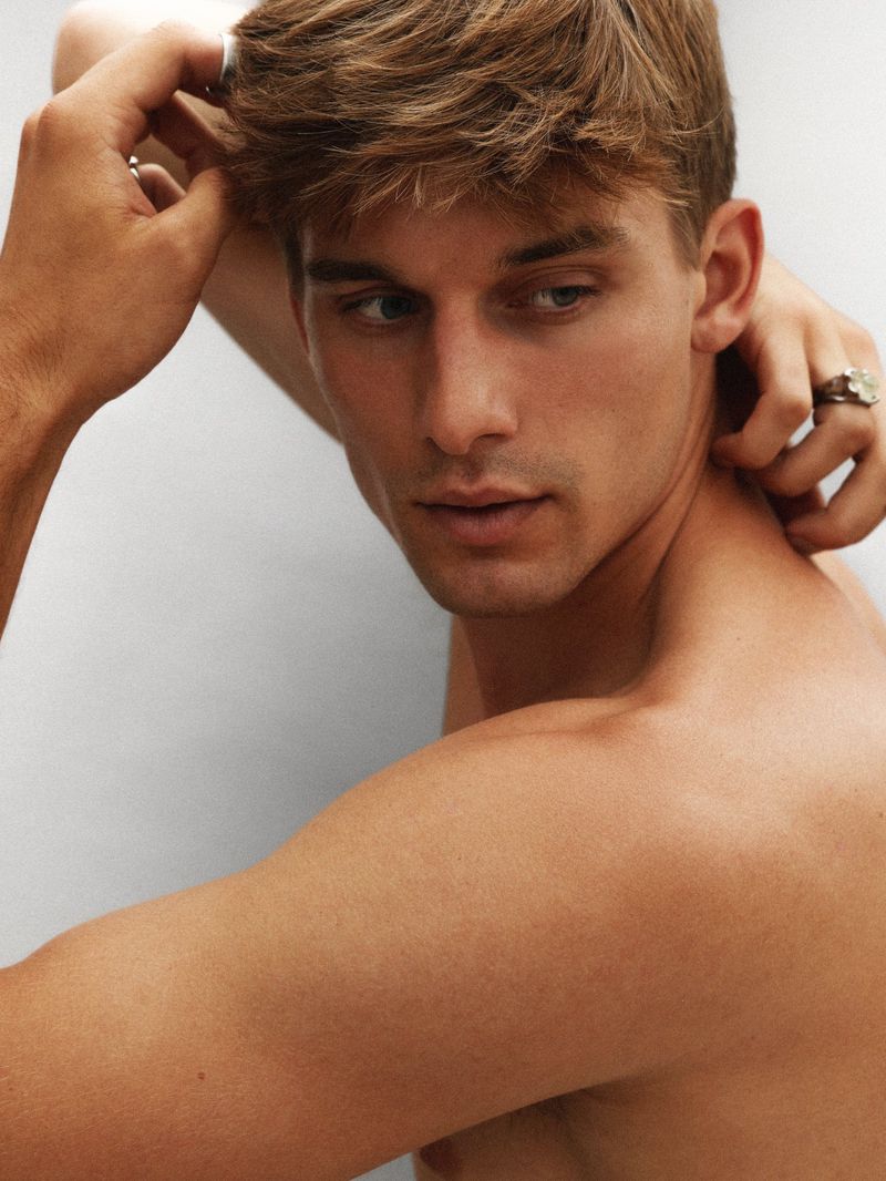 Attractive, fair-skinned adult male posing with hand behind head; natural lighting on clean white background; short blonde hair, clean shaven.