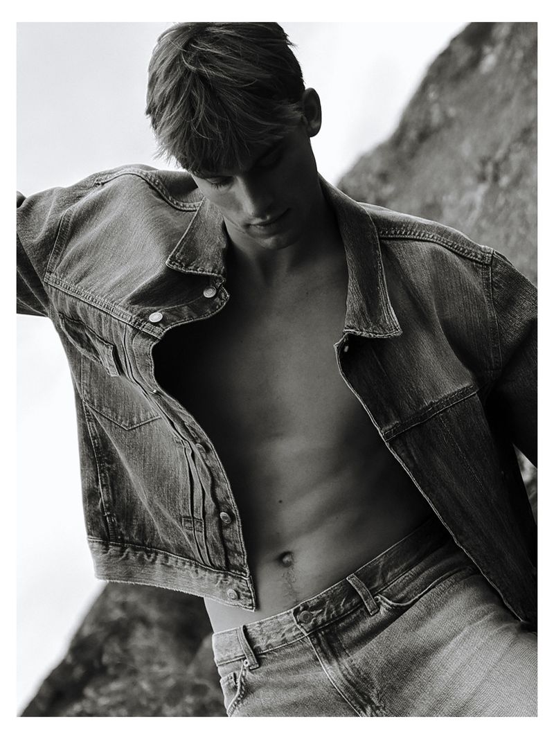Black and white portrait of Mikkel Matthiesen posing shirtless in an open denim jacket and jeans against rocks.