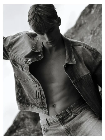 Black and white portrait of Mikkel Matthiesen posing shirtless in an open denim jacket and jeans against rocks.