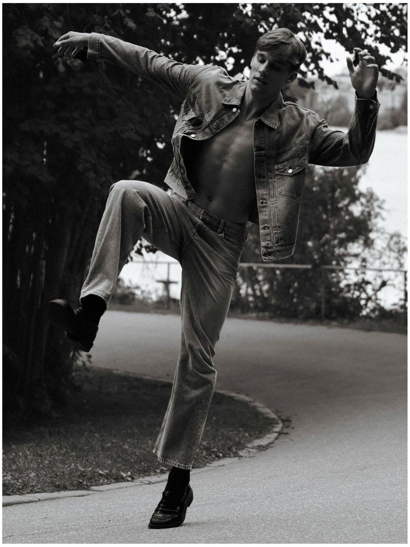 Monochrome fashion shot of a shirtless man in a denim jacket and jeans posing outdoors on a paved path with trees and water in the distance.
