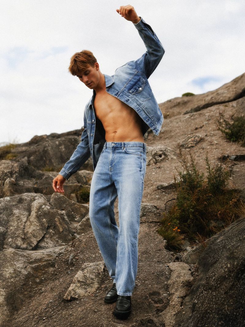 A male model stands on rocks wearing a denim jacket, jeans, and black shoes, posing with one arm up against a cloudy sky backdrop.