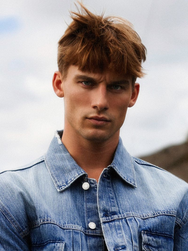 Close-up of a redhead man with a serious expression wearing a denim jacket in an outdoor setting against a cloudy sky backdrop.